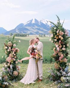 a bride and groom kissing in front of an arch with flowers on the grass, surrounded by mountains