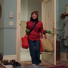 a woman in a red jacket is holding shopping bags and smiling at the camera as she walks into her home