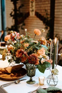 an arrangement of flowers in a vase on top of a table with candles and place settings