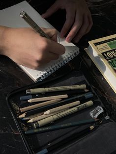 a person is writing on a notebook with several pencils in front of it and an open book next to them