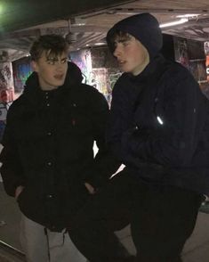 two young men standing next to each other in front of graffiti covered walls at night