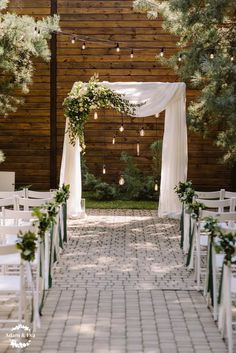 an outdoor ceremony setup with white chairs and greenery on the aisle, surrounded by string lights