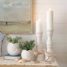 two white candles are sitting on a table next to some seashells and plants