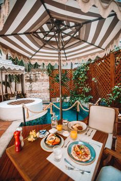 an outdoor dining table with plates and drinks under an umbrella next to a pool in the backyard