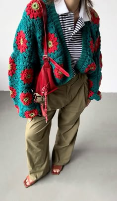 a woman is standing with her hands in her pockets and holding a red purse while wearing an oversize crocheted jacket