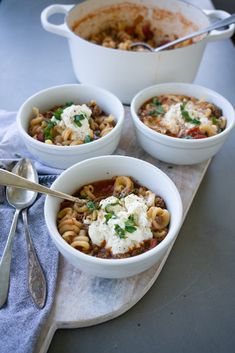 three white bowls filled with pasta and cheese on top of a wooden cutting board next to silver spoons