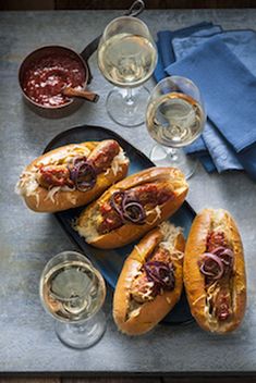 three hot dogs on buns with condiments and glasses of wine next to them