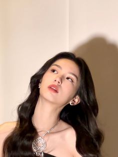 a woman with long black hair wearing a diamond necklace and earrings, standing in front of a white wall