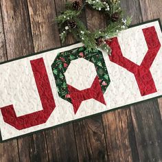 a quilted joy christmas banner hanging on a wooden wall with pine cones and greenery