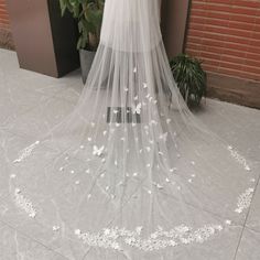 a wedding veil with white butterflies on it sitting next to a potted plant in front of a brick wall