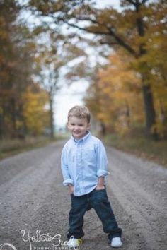 a young boy standing on a dirt road in front of trees with fall foliages