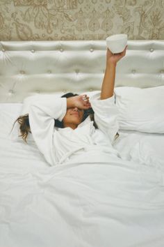 a woman laying in bed with her arms up and holding a cup above her head