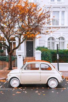 an old car is parked in front of a white house with autumn leaves on the ground