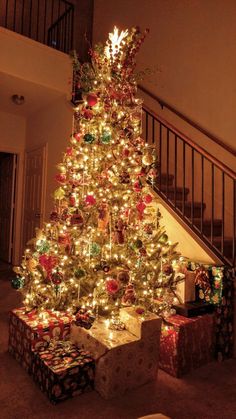 a lit christmas tree with presents under it and stairs in the background at night time