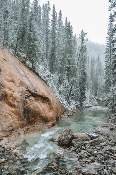 a river running through a forest filled with snow covered rocks and evergreen trees in the background