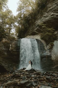 a bride and groom standing in front of a waterfall