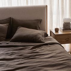 a bed with brown linens and pillows next to a night stand in front of a window