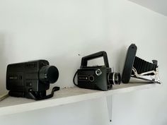 three old cameras sitting on top of a shelf next to each other, one black and one white