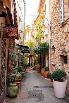 an alleyway with potted plants and tables on either side, surrounded by stone buildings