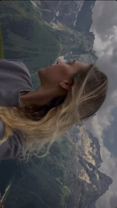 a woman with long blonde hair is looking up at the sky and mountains behind her