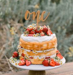 a cake with strawberries and daisies on top sitting on a table next to some bushes