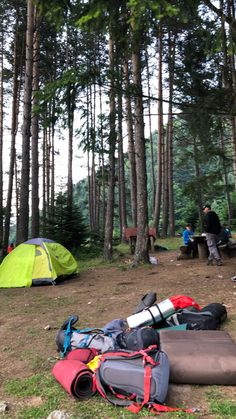 there are many tents in the woods with people sitting at tables behind them and camping equipment on the ground