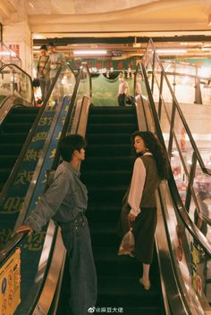 two women are standing on an escalator in a shopping mall, one is holding her hand out to the other