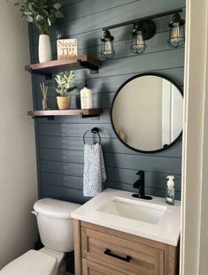 a white toilet sitting next to a bathroom sink under a mirror and wooden shelves above it