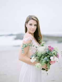 a woman in a white dress holding a bouquet of flowers and greenery on the beach