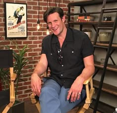 a man sitting in a chair smiling at the camera with a brick wall behind him