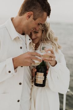 a man and woman standing next to each other holding wine glasses in front of the ocean