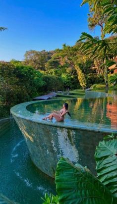 a woman sitting in the middle of a swimming pool surrounded by lush green plants and trees