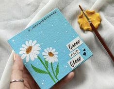 a hand holding a blue notebook with daisies painted on it and an artist's paintbrush next to it