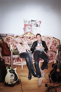 two people sitting on a couch with guitars in front of them and an electric guitar behind them