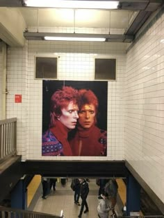 two people standing in front of a large poster on the side of a wall at a subway station