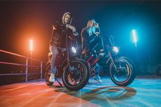 two people standing next to each other on motorcycles in front of some bright colored lights