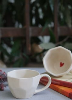 two white cups sitting next to each other on top of a table with books and plants in the background