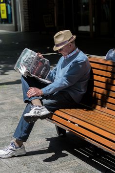 an older man sitting on a wooden bench reading a newspaper while wearing a fedora