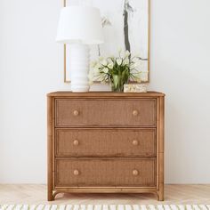 a wooden dresser with flowers on top and a white lamp next to it in a room