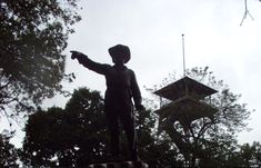 a statue of a man pointing at something in the air with trees behind him and a building in the background