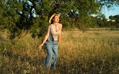 a woman wearing overalls and a straw hat walking through tall grass with trees in the background