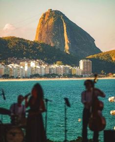 people are playing instruments in front of the water and mountains with buildings in the background