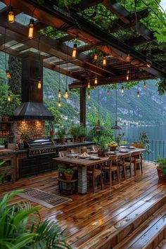 an outdoor kitchen and dining area on a deck overlooking the water with lights hanging from the ceiling