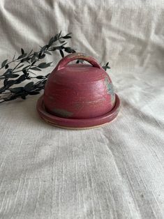 a red tea pot sitting on top of a white bed next to a twig