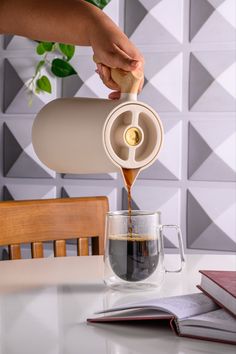 a person pouring coffee into a cup on top of a white table next to a book