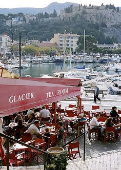 people are sitting at red tables in front of the water with boats docked behind them