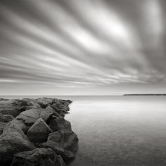 a black and white photo of the ocean with rocks in the foreground, under a cloudy sky