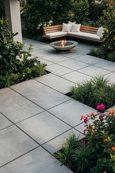 Modern patio with concrete pavers, fire pit, and cushioned bench, surrounded by greenery and flowers. Brushed Concrete Patio Ideas, Outdoor Concrete Patio Ideas, Simple Concrete Patio, Cement Backyard, Stamped Concrete Patio With Seating Wall, Backyard Concrete Slab Ideas, Stamped Concrete Patio Shapes, Cozy Backyard Ideas, Minimalist Patio