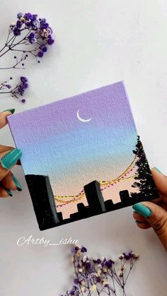 two hands holding up a small card with the city skyline painted on it and purple flowers in the background