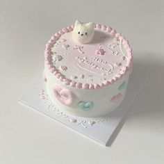 a white cake with pink frosting and a cat decoration on top, sitting on a card